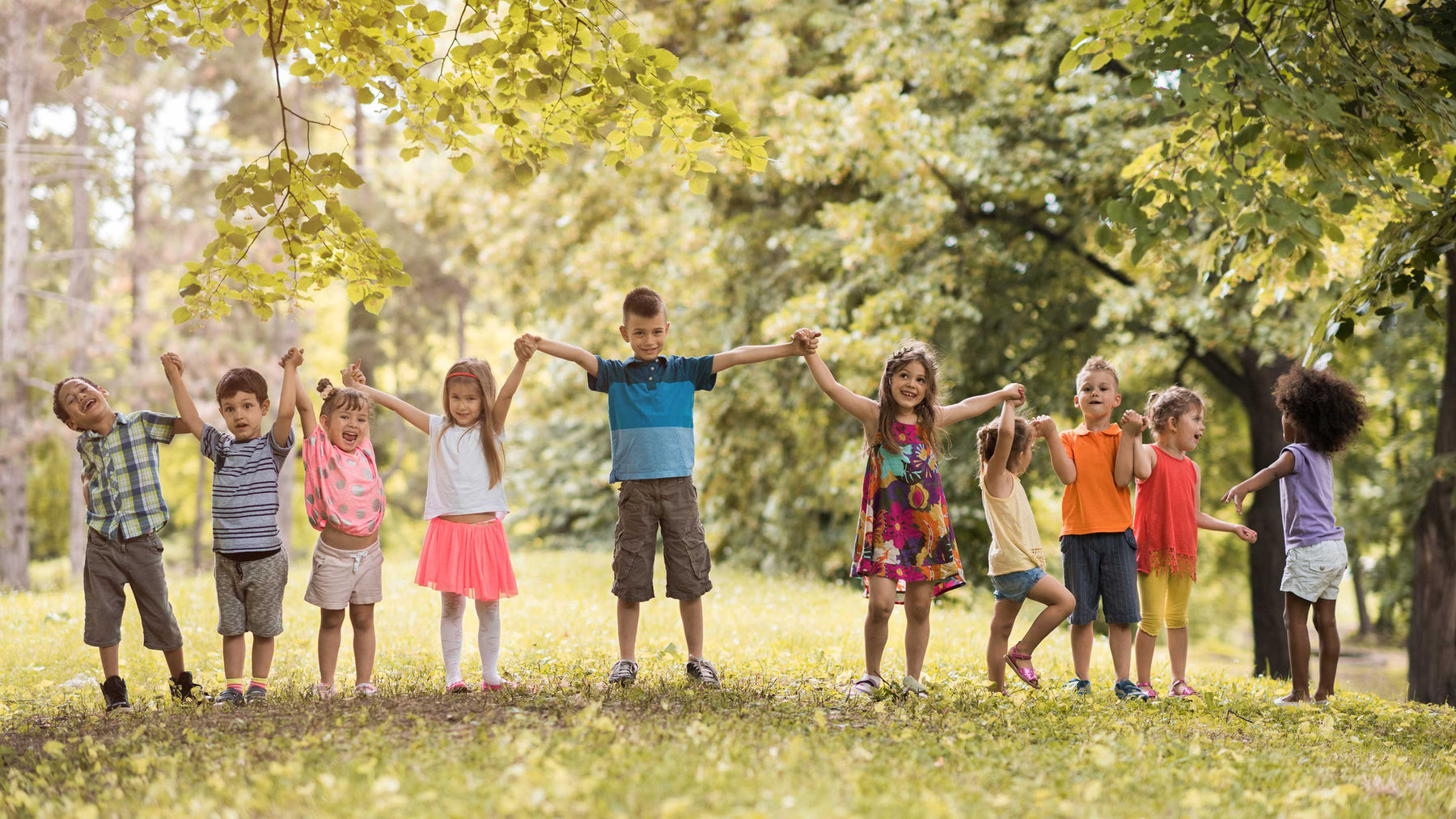 A group of children standing outside in a park in the sunshine holding hands and laughing